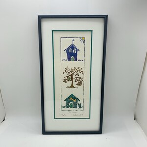 May include: Framed artwork featuring three whimsical scenes. The top panel shows a blue church with a cross and sun. The middle panel depicts a tree with a squirrel. The bottom panel shows a green dog house with a dog. The artwork is framed in a dark blue frame.