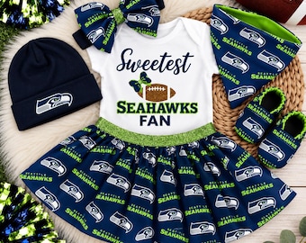 May include: A Seattle Seahawks baby outfit set including a white bodysuit with the text Sweetest Seahawks Fan, a navy blue beanie, a skirt with a green glitter waistband, a hair bow, a bandana bib, and two soft booties, all featuring team logos.
