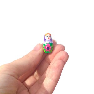 May include: A small, hand-painted wooden nesting doll with a purple and green dress, a pink flower, and a smiling face.