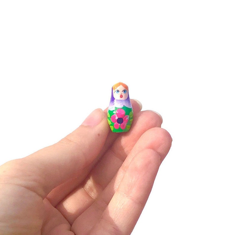 May include: A small, hand-painted wooden nesting doll with a purple and green dress, a pink flower, and a smiling face.