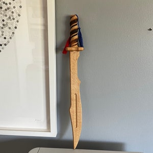 May include: A wooden toy sword with a light brown blade and a dark brown, textured handle. The handle is wrapped with a red and blue tassel. The sword is hanging on a gray wall.