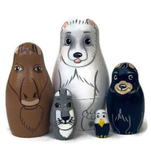 Può includere: Set di cinque bambole russe in legno dipinte a mano, o bambole matrioska, che rappresentano diversi animali. La bambola più grande è un orso polare bianco, seguita da un cavallo marrone, un lupo grigio, un orso nero e un'aquila gialla e nera.