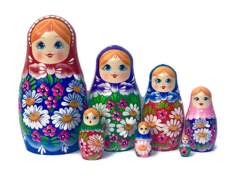 May include: A set of seven traditional Russian nesting dolls, or matryoshka dolls. The dolls are painted with colorful floral patterns and have smiling faces. The largest doll is blue with white flowers, the next is green with white flowers, the next is red with white flowers, the next is pink with white flowers, and the smallest three dolls are red, blue, and green.