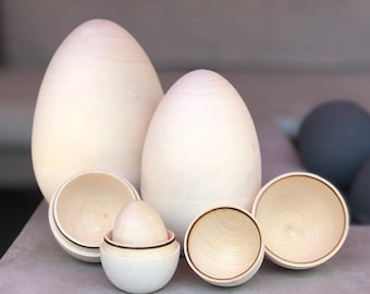 May include: A collection of unfinished wooden egg-shaped nesting dolls, some open to reveal smaller eggs inside. The natural wood grain is visible, and the eggs vary in size. The background is a neutral gray.