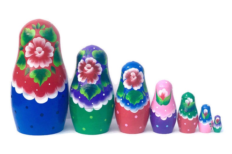 May include: A set of six nesting dolls, also known as Russian nesting dolls, with colorful floral designs. The dolls are painted in shades of blue, red, green, pink, and purple. The largest doll is blue with red and white flowers, the second largest is green with purple and white flowers, the third largest is red with blue and white flowers, the fourth largest is purple with pink and white flowers, the fifth largest is red with white flowers, and the smallest is blue with white flowers.