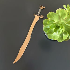 May include: A wooden sword with a curved blade and a detailed handle. The handle features a metal coil and a crossguard. The sword is made of light-colored wood and is set against a dark background.