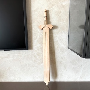Puede incluir: Una espada de madera con acabado marrón claro. El mango tiene un diseño cilíndrico con rayas marrones oscuras. La guarda es de forma simple y angular, y la hoja es larga y recta. La espada está colgada en una pared.