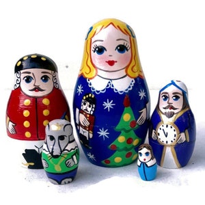May include: A set of five wooden nesting dolls, or matryoshka dolls, depicting characters from the Nutcracker story. The largest doll is a girl with blonde hair and a blue dress, holding a Christmas tree. The other dolls include a nutcracker soldier, a mouse, a man with a clock, and a smaller girl.