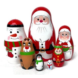 May include: A set of seven hand-painted wooden nesting dolls, or matryoshka dolls, featuring a Christmas theme. The largest doll is Santa Claus, followed by a snowman, a reindeer, a penguin, a girl wearing a red and green elf costume, a woman wearing a red and white dress, and a small snowman.