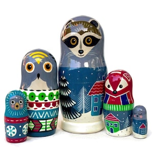 May include: Set of five hand-painted wooden nesting dolls, or matryoshka dolls, featuring winter woodland animals. The largest doll is a raccoon with a blue and white winter scene background. The other dolls are an owl, a fox, and two smaller owls.