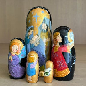 May include: A set of five hand-painted wooden nesting dolls depicting the Nativity scene. The largest doll features a gold background with a star and the figures of Mary, Joseph, and baby Jesus. The other dolls depict angels and a dove.