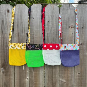 May include: Four mesh crossbody bags hanging on a wooden fence. Each bag has a different color mesh body: yellow, green, white, and purple. The straps and top edges feature various patterned fabrics, including sunflowers, polka dots, and ice cream.