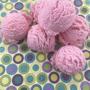 Pode incluir: Cinco bombas de banho cor-de-rosa em forma de cones de gelado, dispostas sobre um fundo colorido de círculos verdes, amarelos, azuis e roxos.