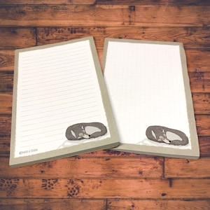 May include: Two lined paper notepads with a sleeping fox illustration at the bottom. The notepads have a light brown border and are on a wooden surface.