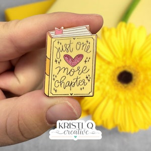 May include: A yellow book-shaped enamel pin with a pink heart and the text "just one more chapter".