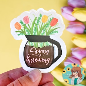 May include: A white sticker with a brown coffee pot filled with pink, yellow, and orange tulips. The text "Spring is brewing" is written in pink on the coffee pot.