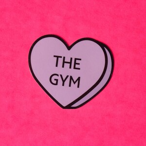 Może przedstawiać: Fioletowa naklejka w kształcie serca z tekstem "THE GYM" na r&oacute;żowym tle.