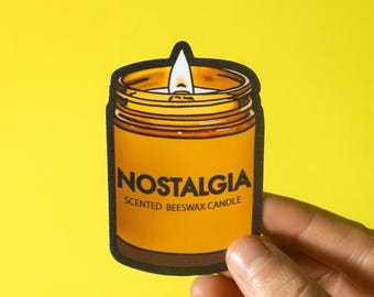 May include: A sticker of a jar-shaped candle with the word "NOSTALGIA" in white text. The candle is orange with a black outline, and the text "SCENTED BEESWAX CANDLE" is below the main word. The background is yellow.