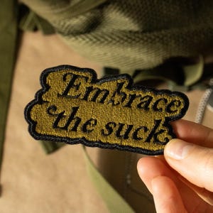 May include: Embroidered patch with the phrase "Embrace the suck" in black script on a gold background. The patch has a black border and is likely designed to be sewn or ironed onto clothing or accessories. The patch is held in a hand.