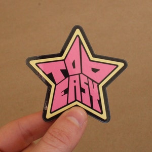May include: A pink and black star-shaped sticker with the text "Too Easy" inside.