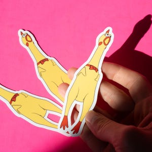 May include: Three yellow rubber chicken stickers with red bow ties. The stickers are on a pink background and are being held by a hand.