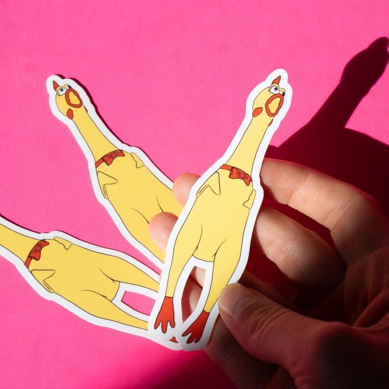 May include: Three yellow rubber chicken stickers with red bow ties. The stickers are on a pink background and are being held by a hand.