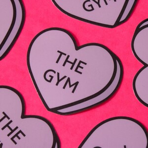 Może przedstawiać: Fioletowe naklejki w kształcie serca z nadrukiem "THE GYM" w kolorze czarnym. Naklejki znajdują się na jasnor&oacute;żowym tle.