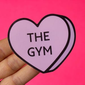 Może przedstawiać: Naklejka w kształcie serca w kolorze fioletowym z czarnym obramowaniem. Naklejka zawiera napis "THE GYM" czarnym kolorem.