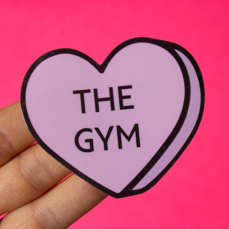 Może przedstawiać: Naklejka w kształcie serca w kolorze fioletowym z czarnym obramowaniem. Naklejka zawiera napis "THE GYM" czarnym kolorem.