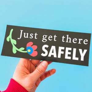 May include: A black rectangular bumper sticker with the words "Just get there SAFELY" in white. A colorful flower and green vine design is on the left side. The sticker is held against a blue background.