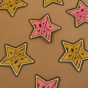 May include: A set of six star-shaped stickers with the text "Too Easy" in a retro font. The stickers are in two colors: pink and gold.