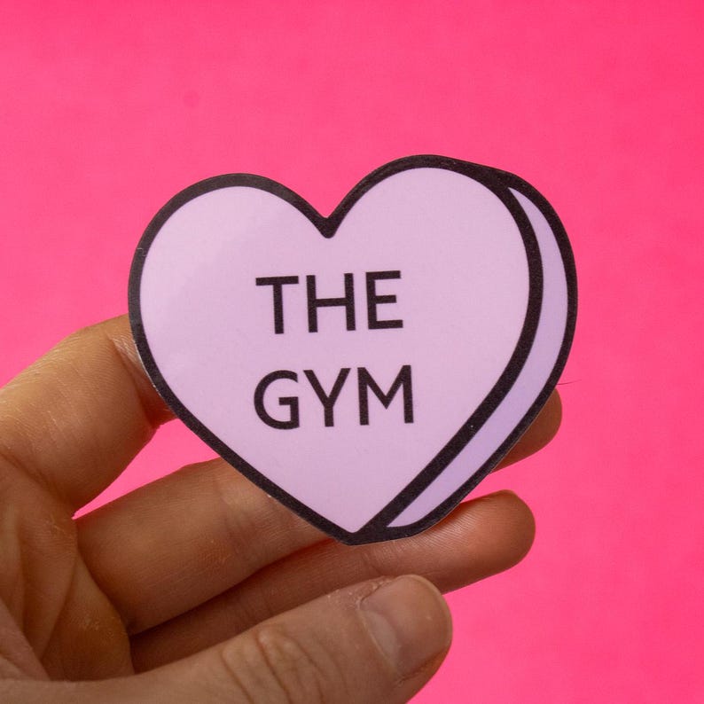 Może przedstawiać: R&oacute;żowa naklejka w kształcie serca z czarnym obramowaniem. Naklejka zawiera napis "THE GYM" czarnym tekstem.