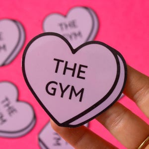 Może przedstawiać: Fioletowa naklejka w kształcie serca z napisem "THE GYM" czarnymi literami. Naklejka znajduje się na r&oacute;żowym tle.