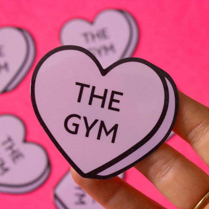 Może przedstawiać: Fioletowa naklejka w kształcie serca z napisem "THE GYM" czarnymi literami. Naklejka znajduje się na r&oacute;żowym tle.