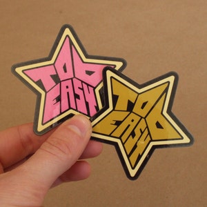 May include: Two star-shaped stickers with the text "Too Easy" in a retro font. One sticker is pink and the other is gold.