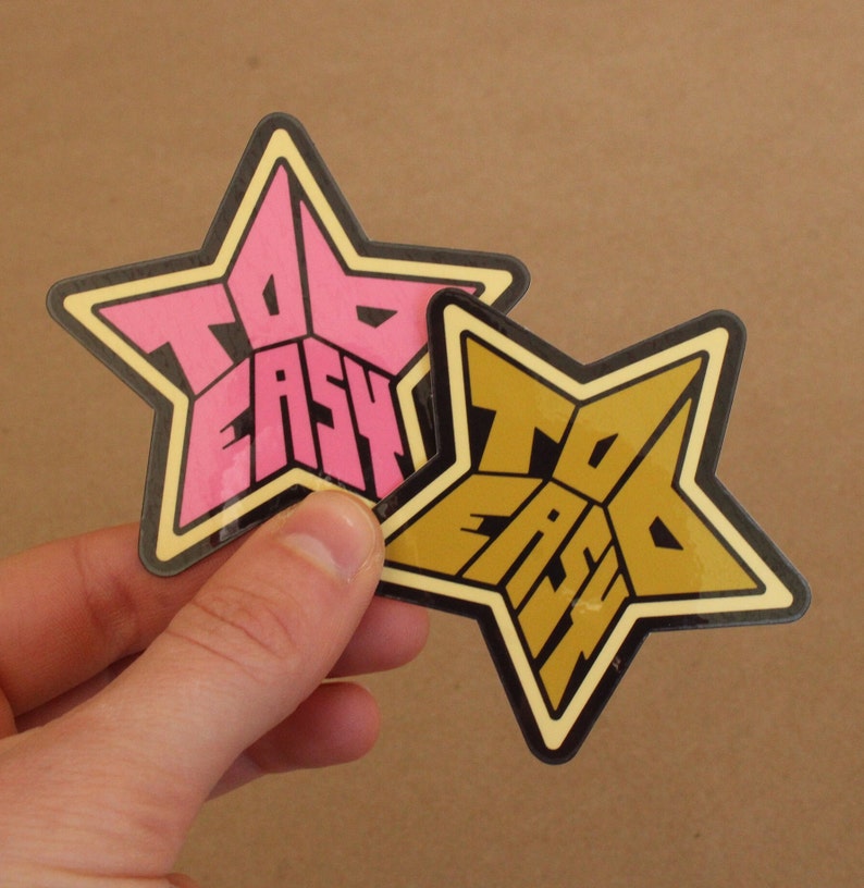 May include: Two star-shaped stickers with the text "Too Easy" in a retro font. One sticker is pink and the other is gold.