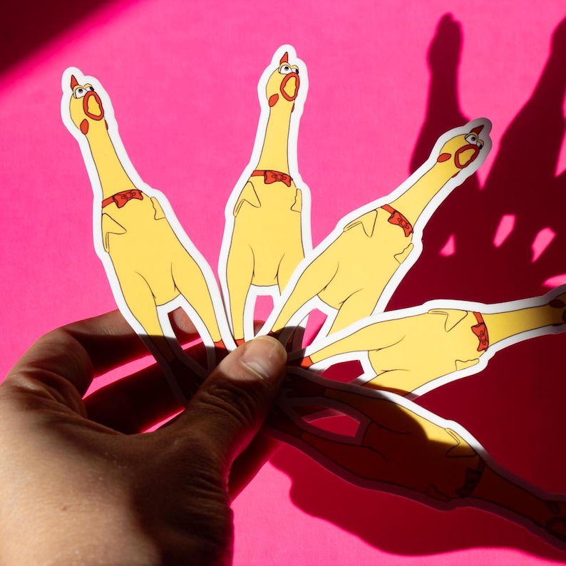 May include: Five yellow rubber chicken stickers with red bow ties. The stickers are held in a hand against a pink background.