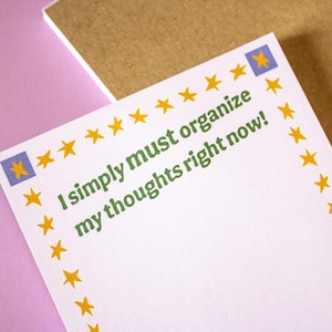 May include: White notepad with the text "I simply must organize my thoughts right now!" in green. The notepad is decorated with yellow stars and blue and yellow star corner accents. The notepad is on a pink surface.