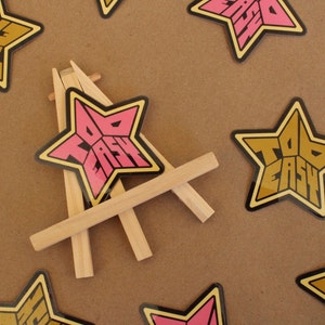 May include: Two star-shaped stickers with the text "Too Easy" in pink and gold letters on a brown background. The pink sticker is on a small wooden easel.