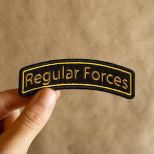 May include: A black embroidered patch with the text "Regular Forces" in gold. The patch has a yellow border and is held by a hand. The background is a neutral colour. The patch is curved.