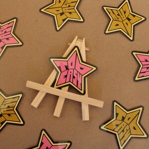 May include: A pink star-shaped sticker with the text "Too Easy" on a wooden easel. The sticker is surrounded by other star-shaped stickers with the same text in gold.