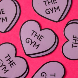 Może przedstawiać: Sześć fioletowych naklejek w kształcie serca z czarnymi obramowaniami i tekstem "THE GYM" na każdej.