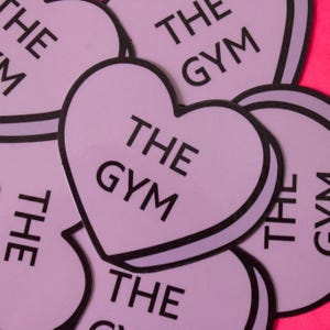 Może przedstawiać: Zbliżenie na kilka r&oacute;żowych naklejek w kształcie serca z nadrukiem "THE GYM" w kolorze czarnym.