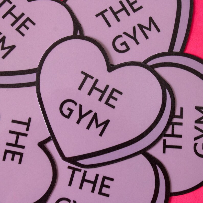 Może przedstawiać: Zbliżenie na kilka r&oacute;żowych naklejek w kształcie serca z nadrukiem "THE GYM" w kolorze czarnym.