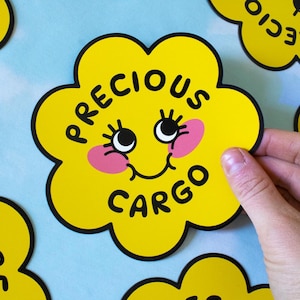 May include: A yellow flower-shaped sticker with a black outline and a smiling face. The sticker says "PRECIOUS CARGO" in black text.
