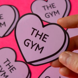 Może przedstawiać: Zestaw fioletowych naklejek w kształcie serca z napisem "THE GYM" na każdej. Naklejki znajdują się na r&oacute;żowym tle.