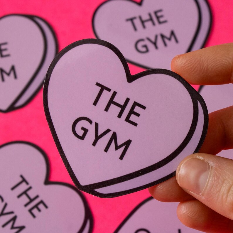 Może przedstawiać: Zestaw fioletowych naklejek w kształcie serca z napisem "THE GYM" na każdej. Naklejki znajdują się na r&oacute;żowym tle.