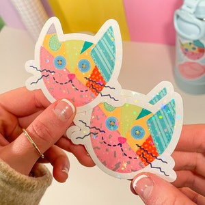 Op de afbeelding: Twee kleurrijke katten stickers met een patchwork ontwerp en glitter accenten. De stickers zijn op een witte achtergrond.