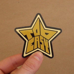May include: A gold star-shaped sticker with the words "Too Easy" written in black letters.