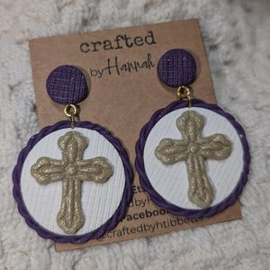 May include: Handmade earrings featuring a gold cross design on a white background, framed by a purple border. The earrings have a matching purple stud top. The earrings are displayed on a brown card with the text "crafted by Hannah".